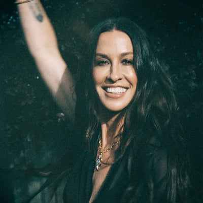 A person with long dark hair, wearing Awe Inspired's Storm Before the Calm Charm Necklace, smiles and raises one arm. The background is dark and leafy, adding to the divine feminine aura she embodies.