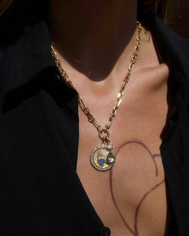 A person wears the Mother Earth Charm Necklace by Awe Inspired, featuring a gold chain with two pendants—a coin-like design and a blue opal gem—against a black shirt.
