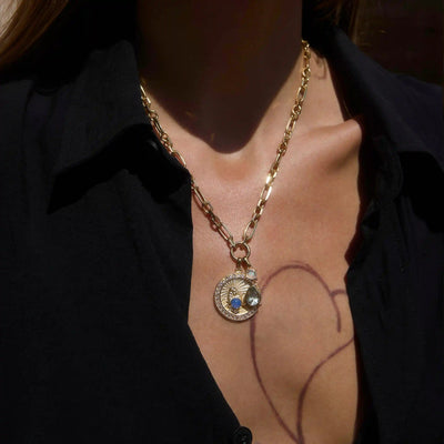 A person wears the Mother Earth Charm Necklace by Awe Inspired, featuring a gold chain with two pendants—a coin-like design and a blue opal gem—against a black shirt.