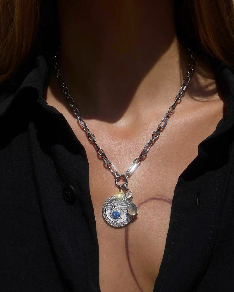 A person wearing the Mother Earth Charm Necklace by Awe Inspired, a silver necklace featuring a circular pendant with a blue opal, paired with a black shirt.
