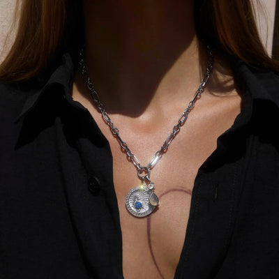 A person wearing the Mother Earth Charm Necklace by Awe Inspired, a silver necklace featuring a circular pendant with a blue opal, paired with a black shirt.