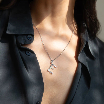 Close-up of a person wearing a partially unbuttoned black shirt, revealing an Awe Inspired Silver Gemstone Initial Necklace with a pendant in the shape of the letter "E".