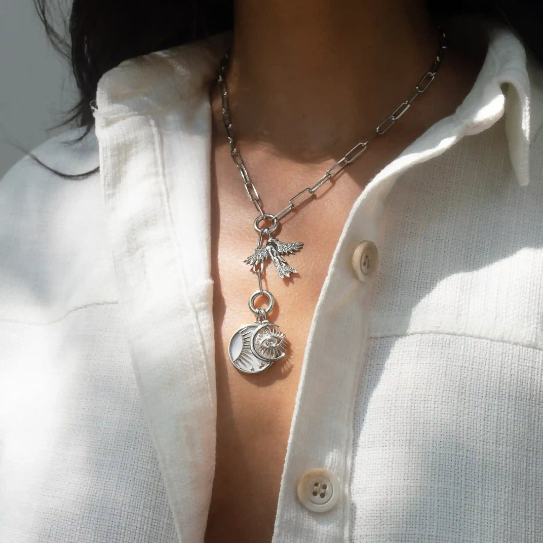 A woman wearing a white buttoned shirt and the Blue Sky Charm Lariat by Awe Inspired, adorned with multiple charm links including a bird, a crescent moon, and a sun.