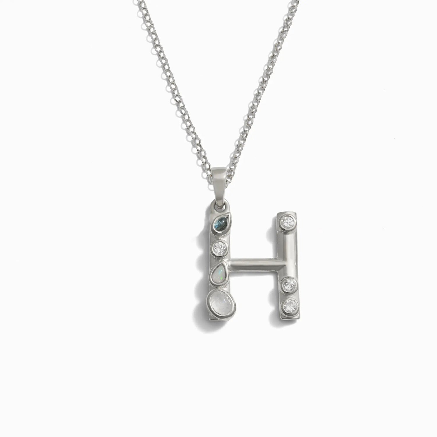 The Silver Gemstone Initial Necklace by Awe Inspired features a pendant in the shape of the letter "H," adorned with bezel set gemstones.