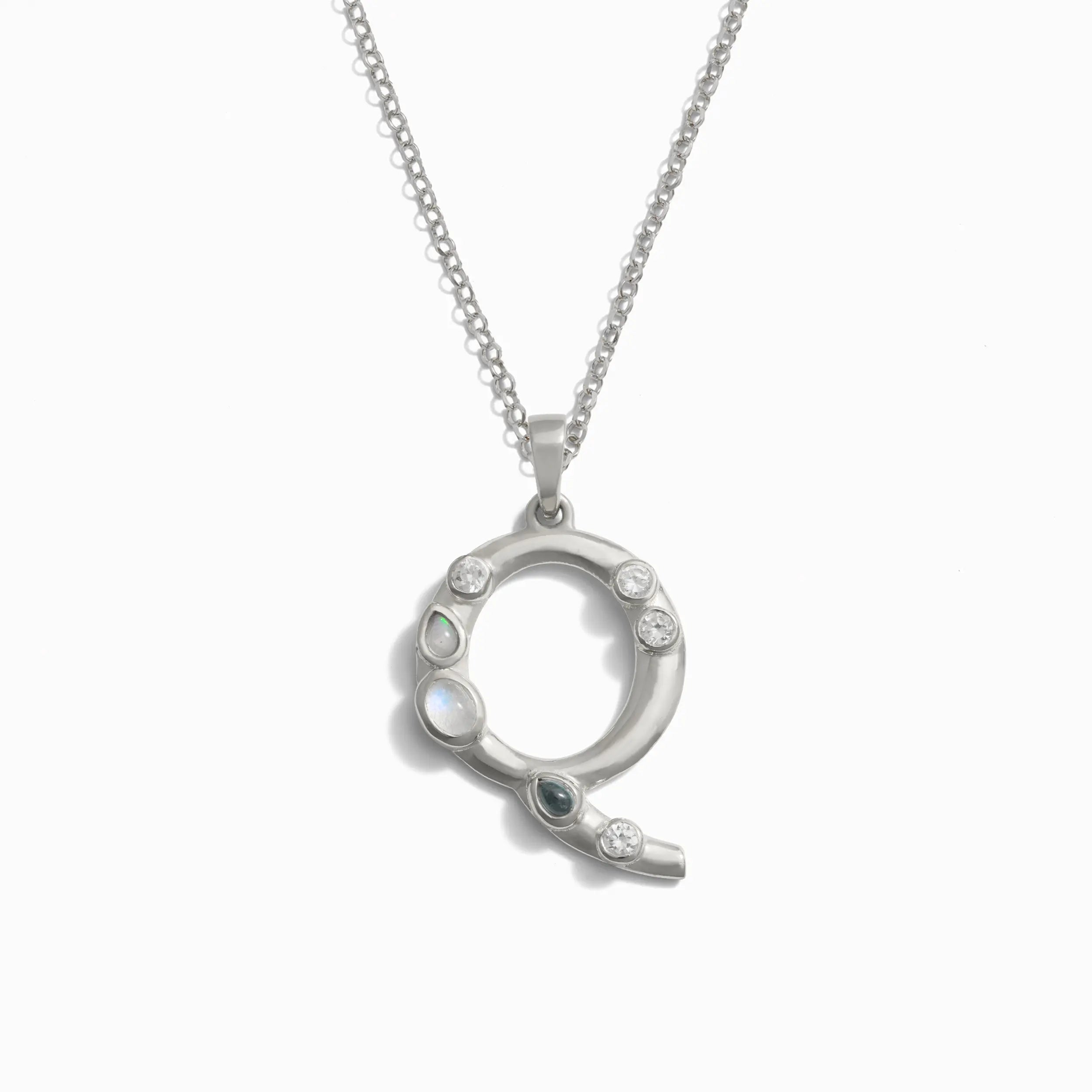 The Silver Gemstone Initial Necklace from Awe Inspired is a silver necklace featuring a statement-size circular pendant adorned with bezel-set gemstones arranged on one side.