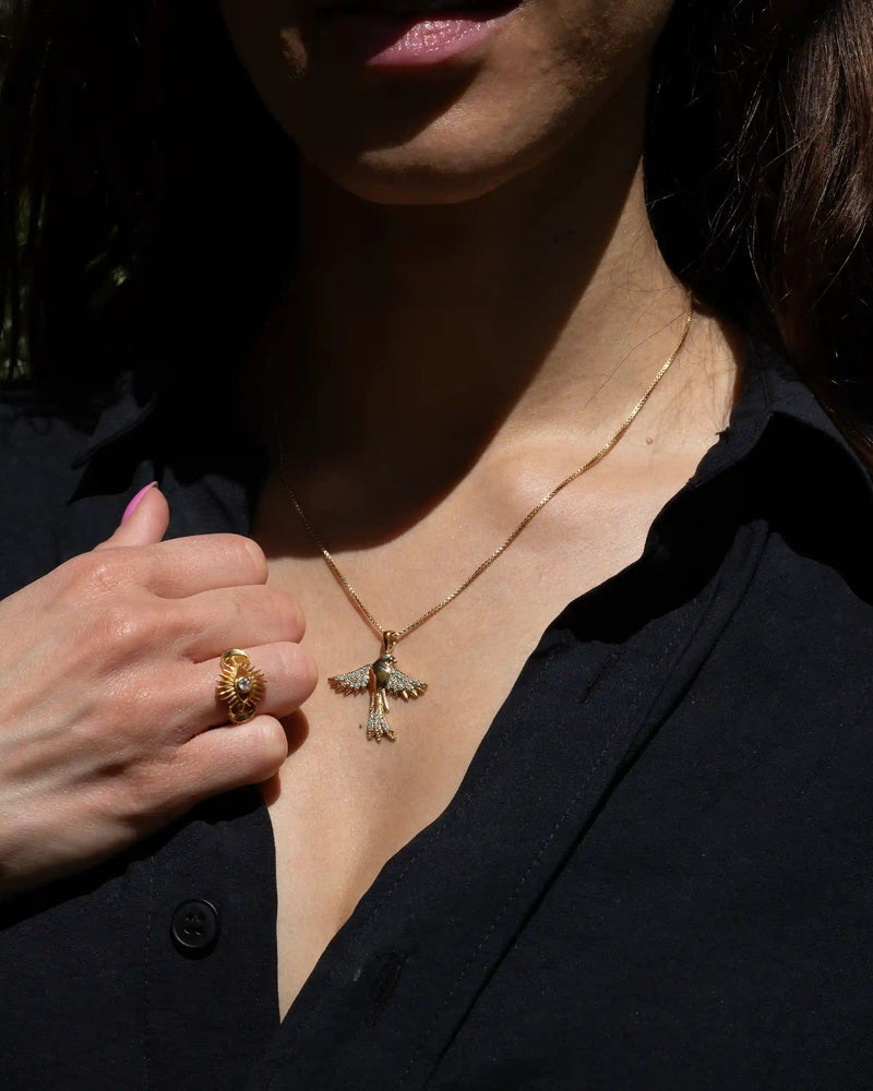 A person wearing a black shirt displays the Awe Inspired Suffragist Bluebird Necklace and a gold ring with an intricate design, accented with blue topaz.