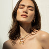 A woman with long brown hair, wearing gold hoop earrings and layered necklaces, including The Charm Collector Necklace by Awe Inspired, stands with closed eyes against a light background.