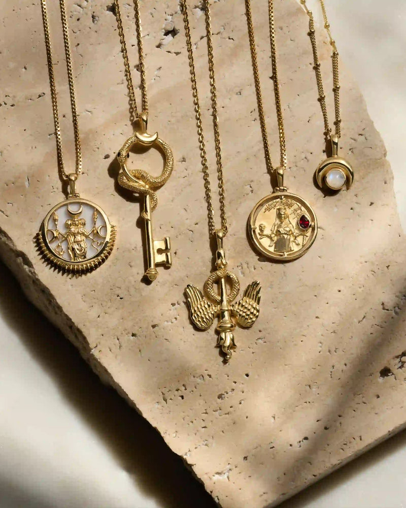 Five gold necklaces with various pendants, including a key, an angel, and round medallions, are arranged on a piece of textured stone. One pendant resembles The Gate of Hades Key Necklace from Awe Inspired, hinting at hidden power and secrets from the Underworld.