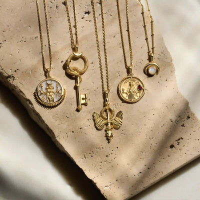 Five gold necklaces with various pendants, including a key, an angel, and round medallions, are arranged on a piece of textured stone. One pendant resembles The Gate of Hades Key Necklace from Awe Inspired, hinting at hidden power and secrets from the Underworld.