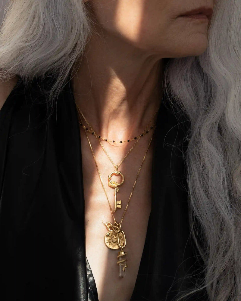 A person with long, gray hair wearing layered gold necklaces, including The Gate of Hades Key Necklace by Awe Inspired and other pendants, paired with a black garment that hints at hidden power.