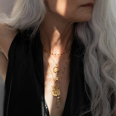 A person with long, gray hair wearing layered gold necklaces, including The Gate of Hades Key Necklace by Awe Inspired and other pendants, paired with a black garment that hints at hidden power.