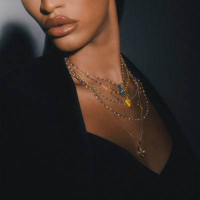 Close-up of a person wearing the Trans Pride Beaded Enamel Necklace by Awe Inspired with colorful triangular pendants and a small round pendant, paired with a dark outfit.