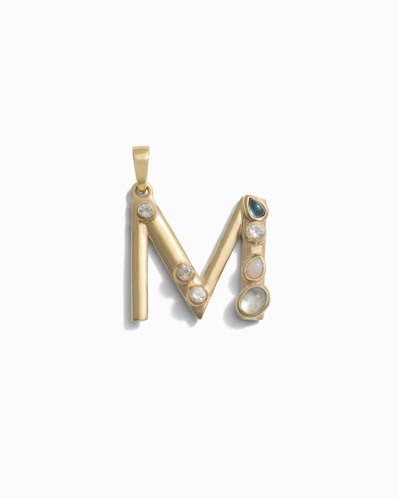 The Awe Inspired Gemstone Initial Pendant features a gold "M" adorned with bezel-set gemstones, including pearls and other stones, all on a white background. This piece of personalized jewelry beautifully showcases initial charms in an elegant and unique design.