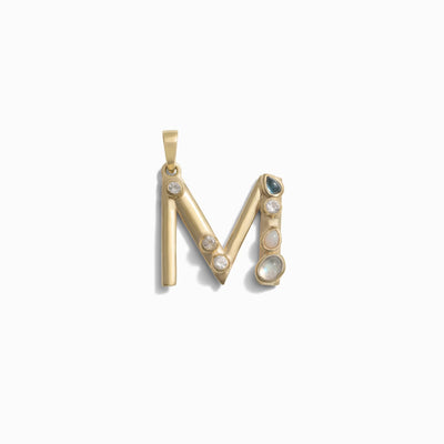 The Awe Inspired Gemstone Initial Pendant features a gold "M" adorned with bezel-set gemstones, including pearls and other stones, all on a white background. This piece of personalized jewelry beautifully showcases initial charms in an elegant and unique design.