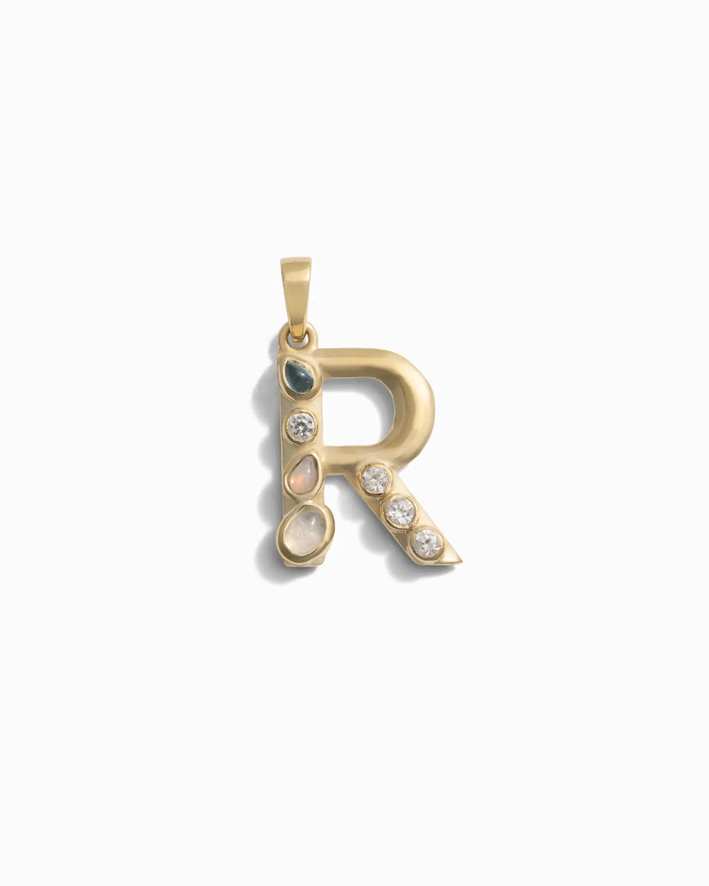 The Awe Inspired Gemstone Initial Pendant is a gold pendant crafted in the shape of the letter "R" and adorned with bezel-set gemstones, including clear, blue, and pink stones. This piece of personalized jewelry makes for a truly unique accessory.