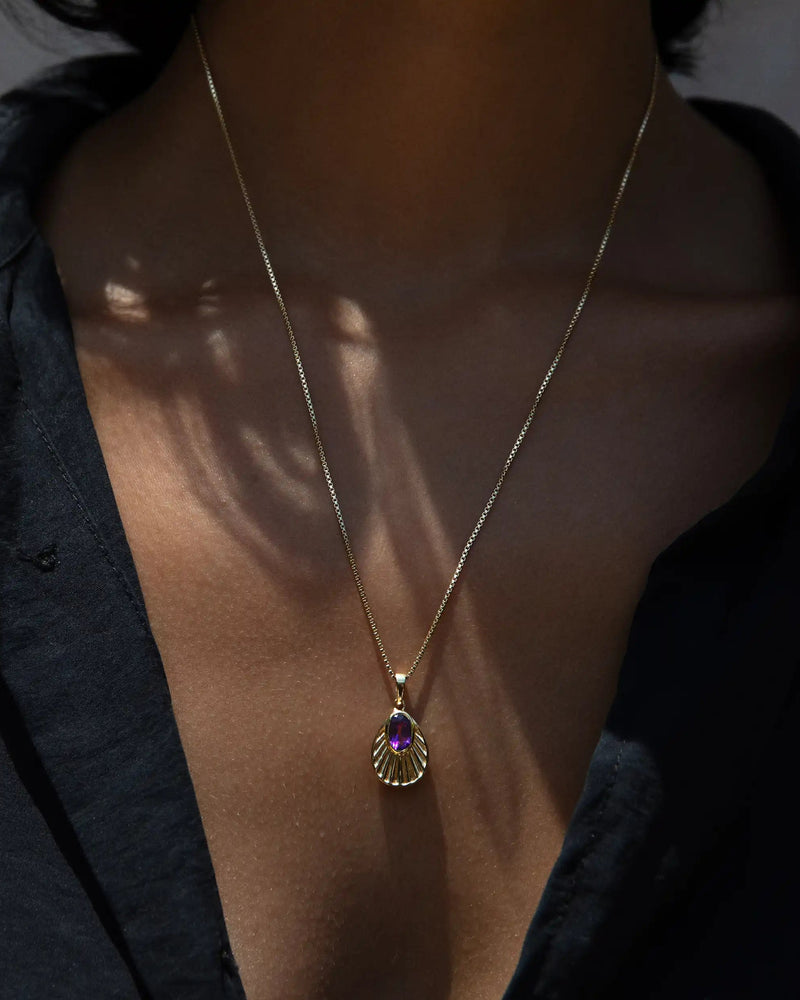 A person wearing the Awe Inspired Aura Pendant radiates an enigmatic aura. The gold necklace with a purple gemstone pendant rests elegantly against their black shirt, adding a touch of mystique to their appearance.