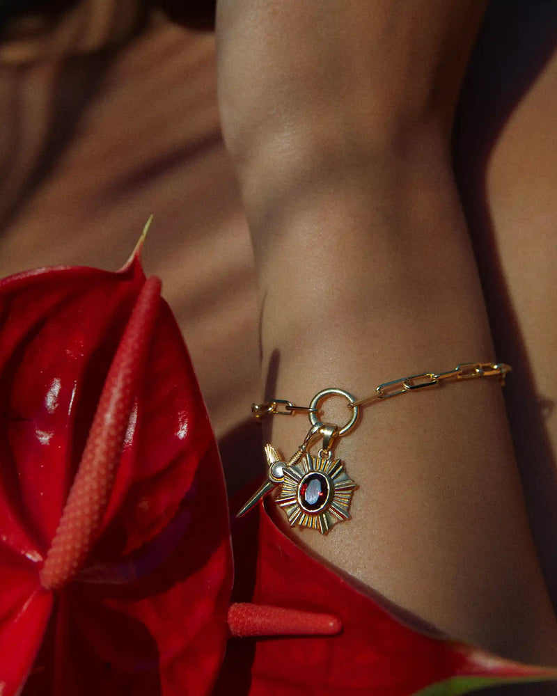 An Aura Pendant by Awe Inspired, featuring a gold bracelet with a sun-shaped pendant adorned with a red gemstone, worn on a wrist surrounded by red tropical leaves, radiating an enchanting aura.