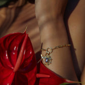 An Aura Pendant by Awe Inspired, featuring a gold bracelet with a sun-shaped pendant adorned with a red gemstone, worn on a wrist surrounded by red tropical leaves, radiating an enchanting aura.