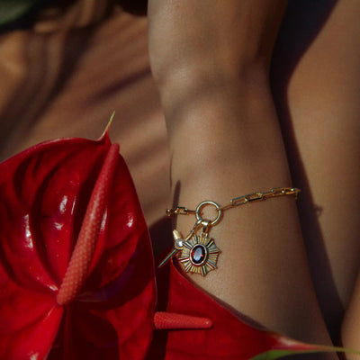 An Aura Pendant by Awe Inspired, featuring a gold bracelet with a sun-shaped pendant adorned with a red gemstone, worn on a wrist surrounded by red tropical leaves, radiating an enchanting aura.