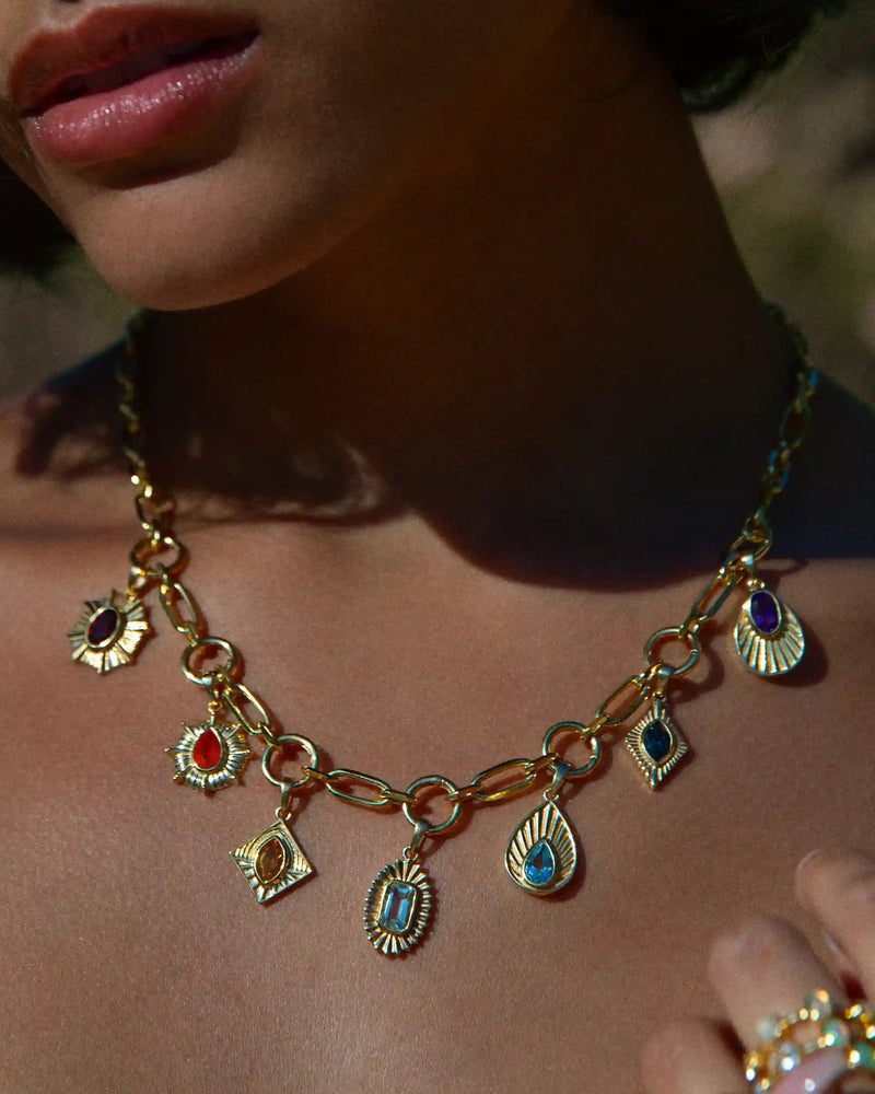 Close-up of a person wearing the Aura Pendant by Awe Inspired, a gold chain necklace adorned with various colorful gemstone chakra pendants. The person is also wearing gold rings, and the background is blurred.