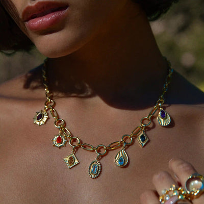 Close-up of a person wearing the Aura Pendant by Awe Inspired, a gold chain necklace adorned with various colorful gemstone chakra pendants. The person is also wearing gold rings, and the background is blurred.