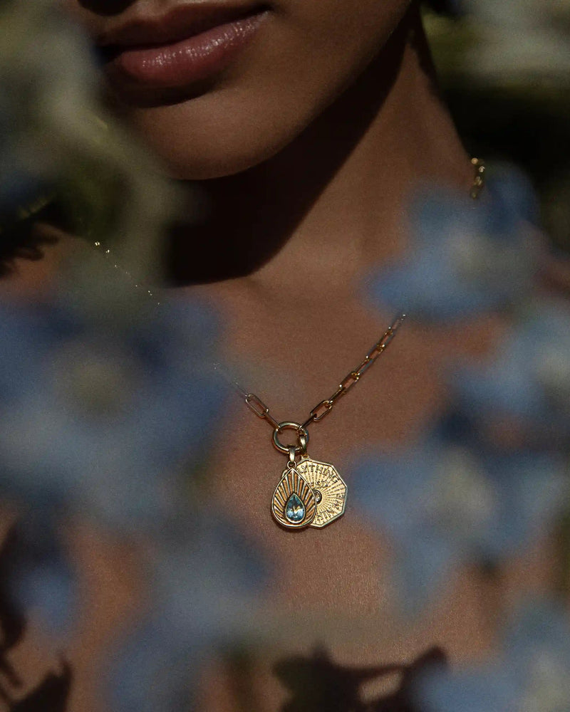 A person adorned with the Awe Inspired Aura Pendant—a gold necklace featuring a shell-shaped pendant and a shimmering blue gemstone—is partially hidden by out-of-focus blue flowers, creating an aura of mystery.