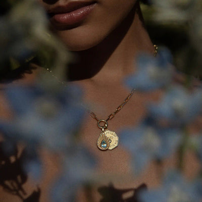 A person adorned with the Awe Inspired Aura Pendant—a gold necklace featuring a shell-shaped pendant and a shimmering blue gemstone—is partially hidden by out-of-focus blue flowers, creating an aura of mystery.