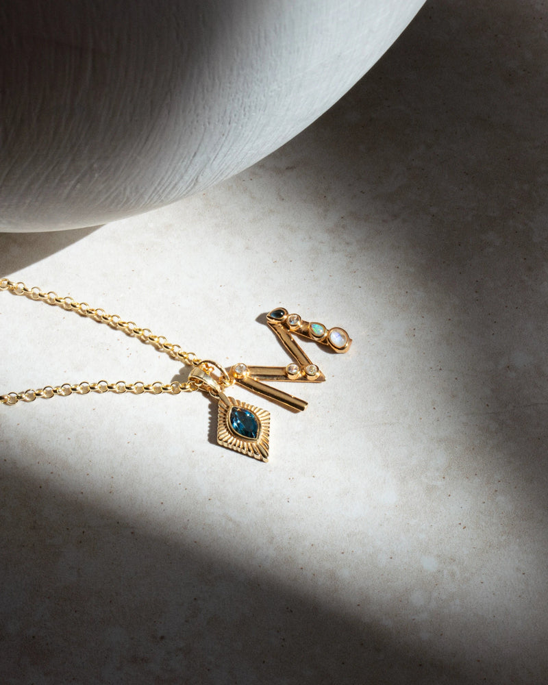 A Gold Gemstone Initial Necklace by Awe Inspired, featuring a blue gemstone bezel set pendant, and a pair of gold earrings adorned with small gemstones, all gracefully displayed on a textured surface.