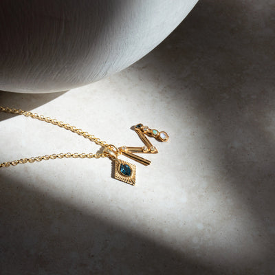 A Gold Gemstone Initial Necklace by Awe Inspired, featuring a blue gemstone bezel set pendant, and a pair of gold earrings adorned with small gemstones, all gracefully displayed on a textured surface.