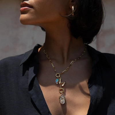 A person wearing a Gemstone Initial Pendant by Awe Inspired, along with multiple other pendants and personalized jewelry, paired with a dark, open-collared shirt.