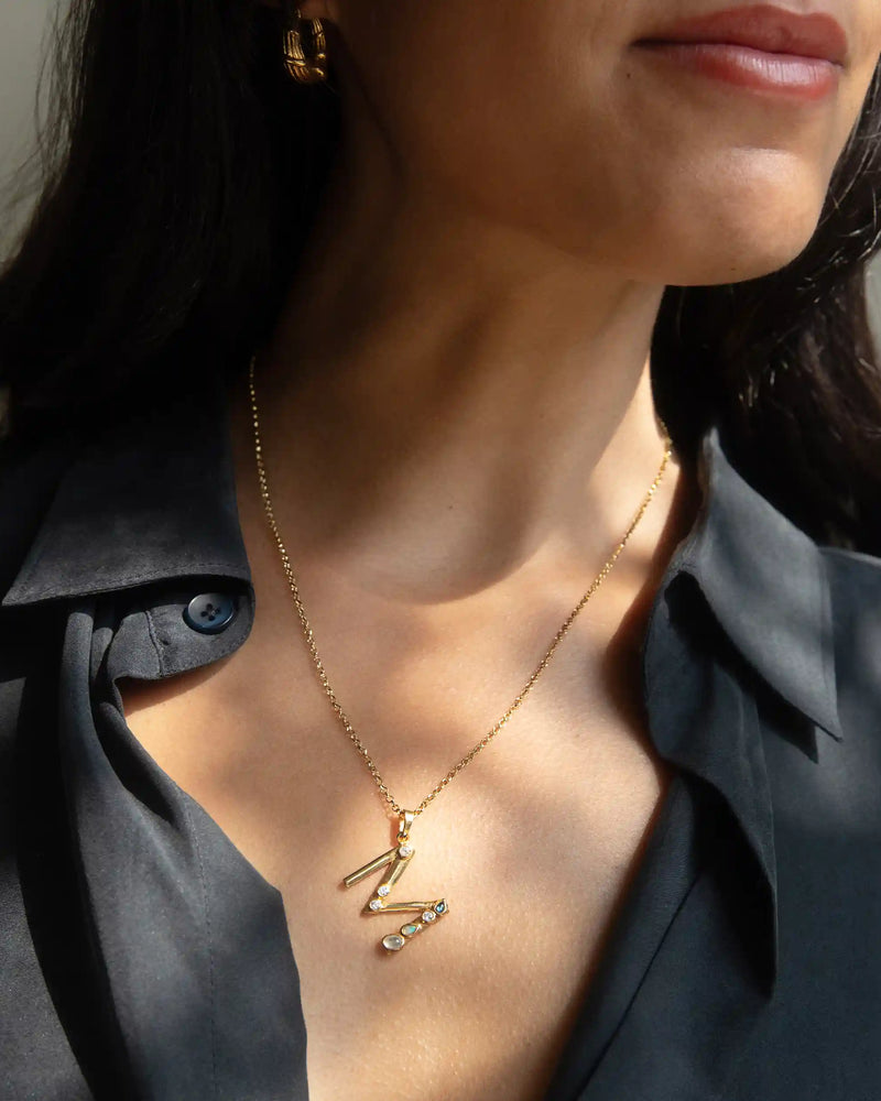 A woman wearing a black blouse showcases the Awe Inspired Gold Gemstone Initial Necklace with a zigzag pendant and bezel-set gemstones, accompanied by gold earrings.