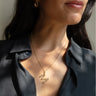 A woman wearing a black blouse showcases the Awe Inspired Gold Gemstone Initial Necklace with a zigzag pendant and bezel-set gemstones, accompanied by gold earrings.