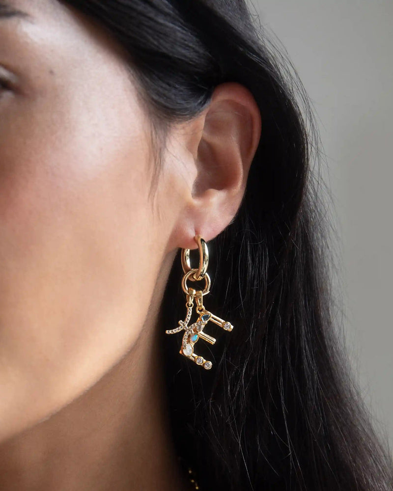 Close-up of a woman's ear adorned with the Awe Inspired Gemstone Initial Pendant, featuring bezel set gemstones. Her face is partially visible, accentuated by her long, dark hair.