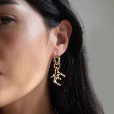 Close-up of a woman's ear adorned with the Awe Inspired Gemstone Initial Pendant, featuring bezel set gemstones. Her face is partially visible, accentuated by her long, dark hair.