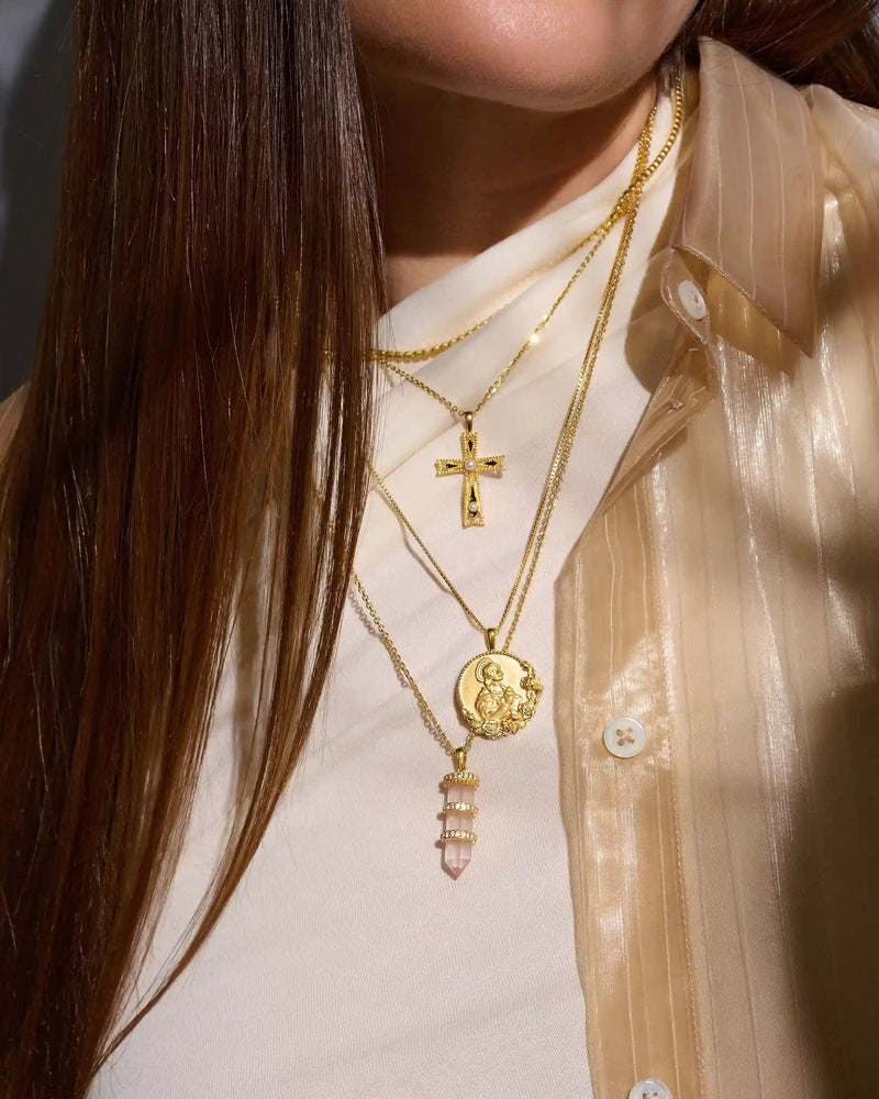 A person wearing a translucent beige shirt and white top, layered with multiple gold necklaces featuring various pendants, including a cross, a medallion, and the Manifestation Crystal Amulet by Awe Inspired.