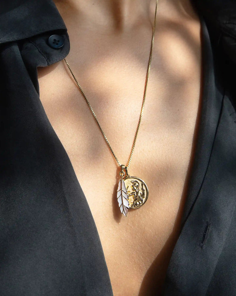 A close-up of a person wearing a black top with Awe Inspired's Mother of Pearl Feather Amulet, featuring a circular pendant and an owl-shaped charm, evoking Athena's power.