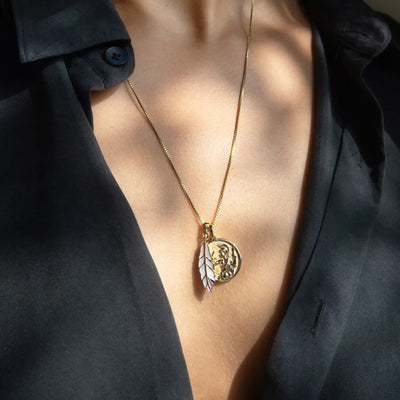 A close-up of a person wearing a black top with Awe Inspired's Mother of Pearl Feather Amulet, featuring a circular pendant and an owl-shaped charm, evoking Athena's power.