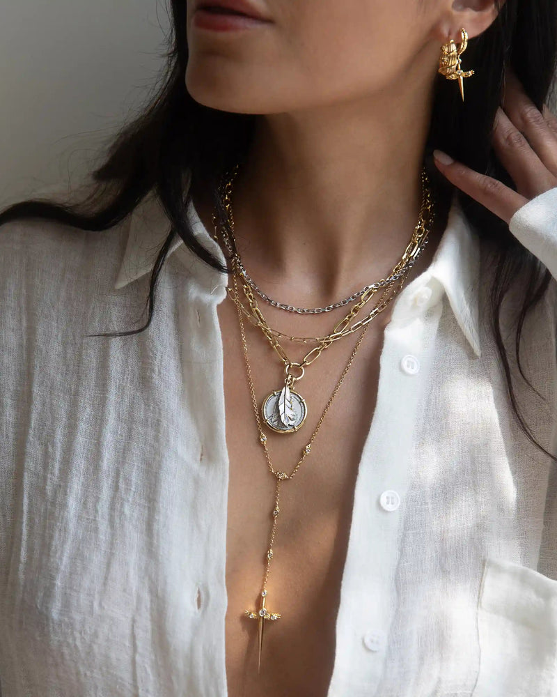 A woman is wearing multiple layered necklaces, including the Awe Inspired Mother of Pearl Feather Amulet, and a cross pendant. She is dressed in a white partially unbuttoned shirt, has gold earrings, and is touching her hair with one hand.