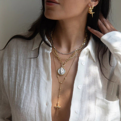 A woman is wearing multiple layered necklaces, including the Awe Inspired Mother of Pearl Feather Amulet, and a cross pendant. She is dressed in a white partially unbuttoned shirt, has gold earrings, and is touching her hair with one hand.