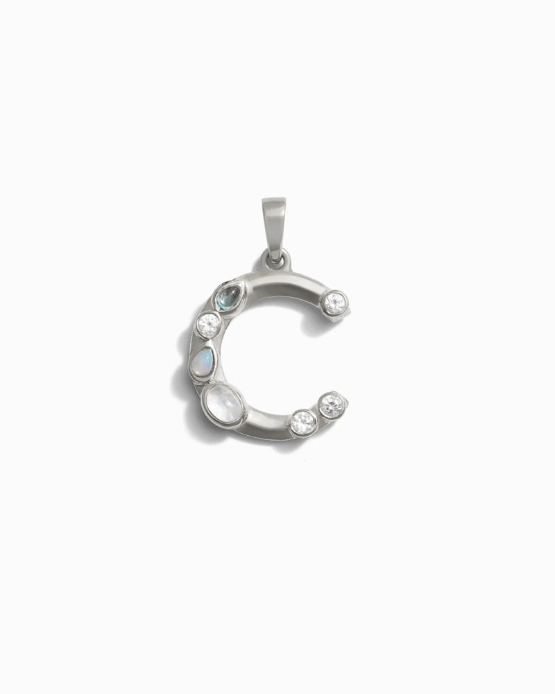 The Awe Inspired Gemstone Initial Pendant features a silver crescent-shaped design adorned with bezel set gemstones in both round and oval cuts, elegantly arranged along its curve.