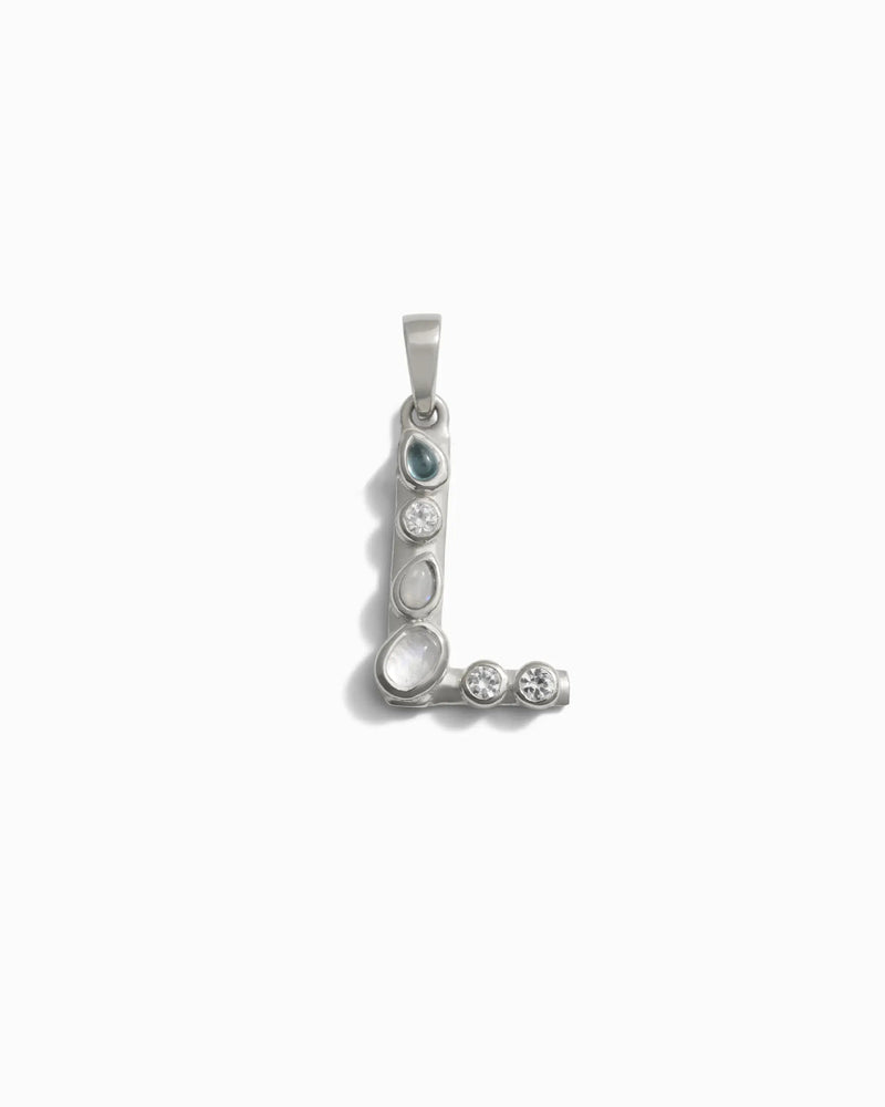 The Gemstone Initial Pendant by Awe Inspired features a silver pendant shaped like the letter "L," adorned with bezel set gemstones along its form, resting elegantly against a white background. This exquisite piece of personalized jewelry is an ideal choice for those who cherish initial charms.