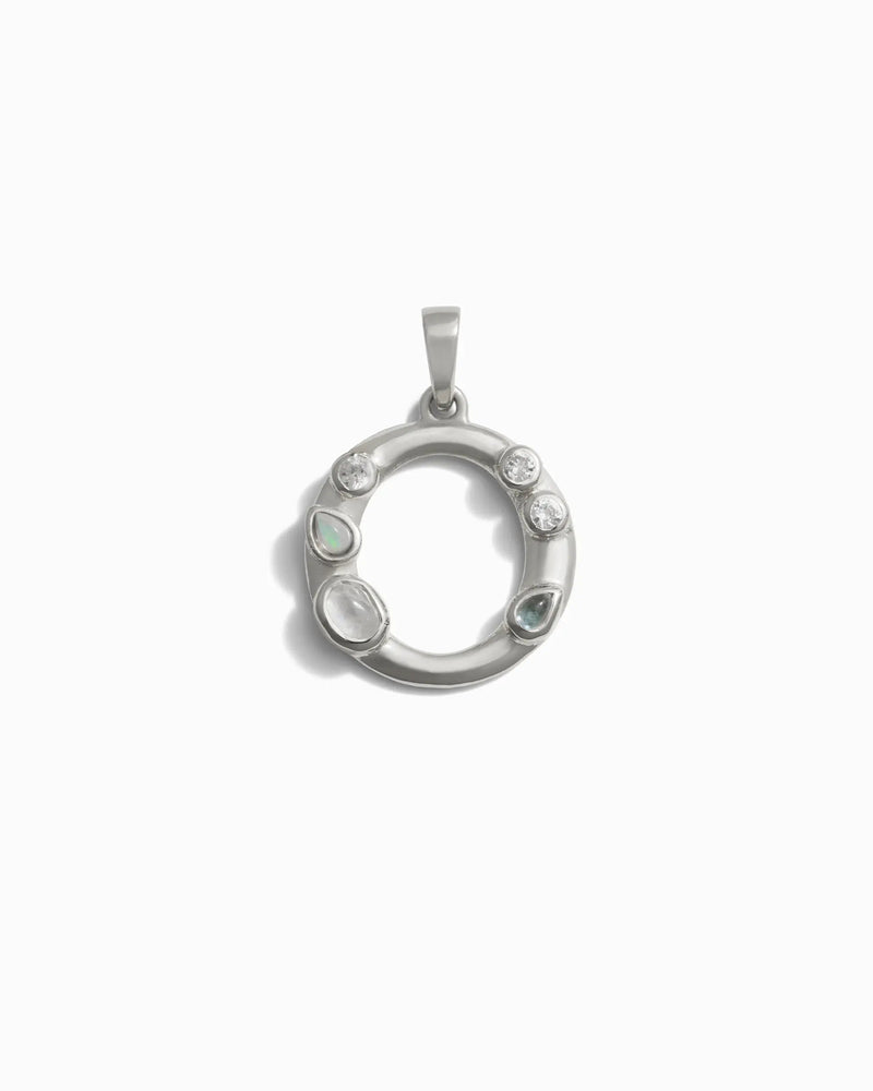 The Gemstone Initial Pendant by Awe Inspired is a circular silver pendant adorned with various bezel-set gemstones, including small circular and teardrop shapes, on a white background.