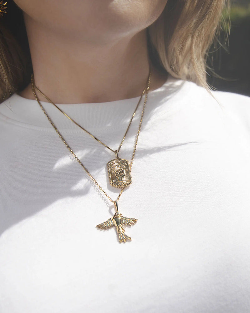 A woman wearing two gold necklaces, one featuring the Suffragist Bluebird Amulet with a blue topaz pendant resembling a bird from Awe Inspired and the other with an engraved rectangular pendant. She is dressed in a white shirt.
