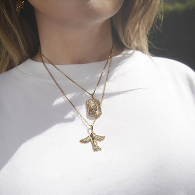 A woman wearing two gold necklaces, one featuring the Suffragist Bluebird Amulet with a blue topaz pendant resembling a bird from Awe Inspired and the other with an engraved rectangular pendant. She is dressed in a white shirt.