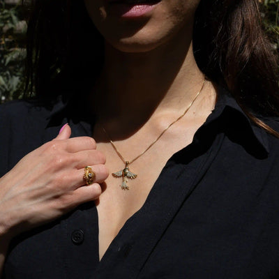 Person wearing a black top holds a hand near their chest, showcasing a gold ring and the Suffragist Bluebird Amulet by Awe Inspired.