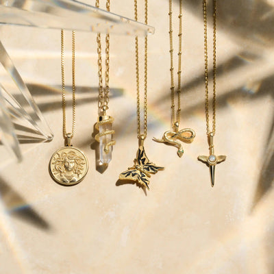 A collection of five gold necklaces displayed on a beige surface, each with a unique pendant: a sun medallion, The Guardian’s Crystal Amulet by Awe Inspired with a snake, butterfly, snake, and a gemstone spear.