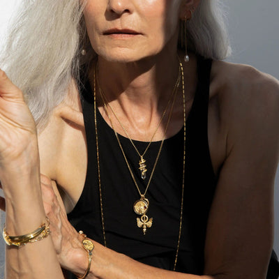 Woman with long white hair wearing multiple gold necklaces and a black sleeveless top. She is also adorned with gold bracelets and earrings, one of which features The Guardian's Crystal Amulet by Awe Inspired, hinting at her afterlife connection.