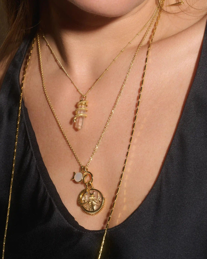 Close-up of a person wearing two gold necklaces. One features The Guardian's Crystal Amulet by Awe Inspired, known to help communicate with the dearly departed, while the other displays a circular pendant with a star design. The person is donning a dark top.