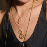 Close-up of a person wearing two gold necklaces. One features The Guardian's Crystal Amulet by Awe Inspired, known to help communicate with the dearly departed, while the other displays a circular pendant with a star design. The person is donning a dark top.