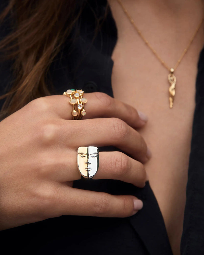 Close-up of a person wearing an Awe Inspired gold necklace and multiple rings. A prominent stack of Face to Face Rings is on their index finger, and a decorative nesting ring is on their pinky finger, over a black garment.